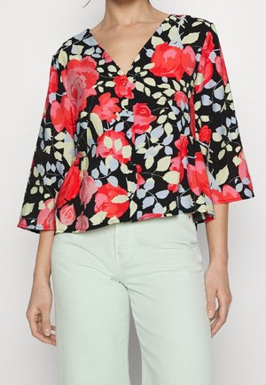 Kvinde iført en sort blomstret bluse med røde, pink og lysegrønne blomster og lyse bukser.