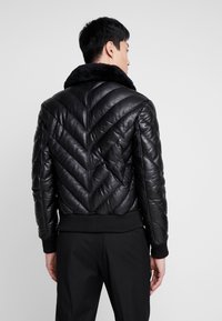 Zwarte leren pufferjack met een chevron-quiltpatroon, voorzien van een schapenwollen kraag en ribgebreide boorden, gezien vanuit een achteraanzicht.