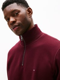 Burgunderfarget halv-zip genser laget av myk stoff, med en liten logo detalj på brystet og en strukturert strikket krage.