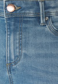 Jeans de mezclilla azul claro con detalles de botones de cobre, costura vertical y una textura desgastada. Diseño clásico de cinco bolsillos visible.
