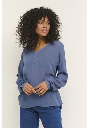 Blusa a maniche lunghe azzurro chiaro con scollatura a V, tessuto texturizzato, vestibilità rilassata e polsini arricciati. Abbinata a jeans blu.