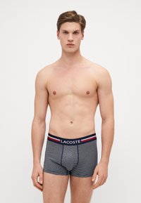 Navy gestreifte Boxershorts mit Lacoste-Logo am Bund. Verfügt über einen elastischen Bund mit roten und weißen Akzenten.