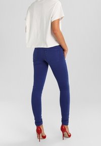 Nainen, joka on pukeutunut valkoiseen lyhythihaiseen paitaan, sinisiin tiukkoihin farkkuihin ja punaisiin korkokenkiin, seisoo yksi käsi taskussa.