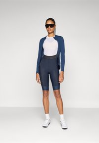 Idrettsantrekk med en marineblå jakke med glidelås, hvit mesh-t-skjorte og marineblå sykkelshorts med svart liv. Solbriller og hvite sko.