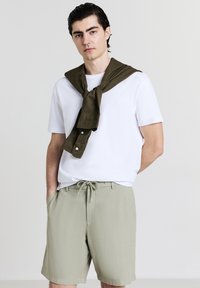 Jeune homme aux cheveux gominés portant un t-shirt blanc, un short kaki avec cordon de serrage et une chemise vert olive drapée sur ses épaules.