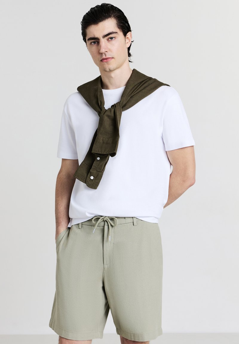 Jeune homme aux cheveux gominés portant un t-shirt blanc, un short kaki avec cordon de serrage et une chemise vert olive drapée sur ses épaules.