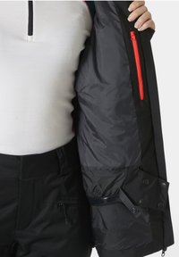 Černá bunda s hladkým podšívkovým interiérem, vybavená jasně červenou zipovou kapsou a nastavitelnými kovovými uzávěry. Pod ní je viditelná bílá základní vrstva.