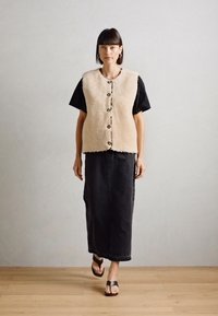 Beige-Fellweste mit runden Knöpfen, getragen über einem schwarzen T-Shirt mit kurzen Ärmeln, kombiniert mit einem langen schwarzen Rock und braunen Sandalen. Glatte Textur.