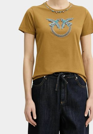 Donna che indossa una maglietta a maniche corte color senape con motivo di uccello turchese e perline, abbinata a jeans scuri in denim annodati in vita.