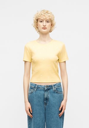 Γυναίκα με σγουρά ξανθά μαλλιά, φορώντας κίτρινη κοντομάνικη μπλούζα με μικρές λευκές βούλες και ψηλόμεσο μπλε τζιν, στέκεται μπροστά σε λευκό φόντο.