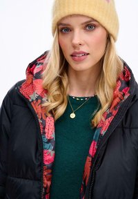 Čierna prešívaná bunda s kvetinovým vzorom vo vnútri, zelený sveter pod ňou a žltá pletená čiapka. Obsahuje zlatý náhrdelník a dlhé, vlnité vlasy.