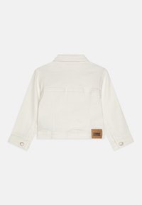 Bijela traper jakna s dugim rukavima, ovratnikom i dugmadima na rukavima. Ima smeđu oznaku s natpisom "TOMMY HILFIGER" na leđima.