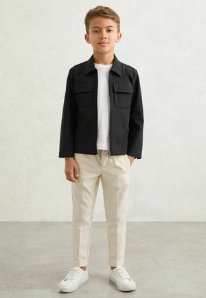 Băiat stând pe un fundal simplu, purtând o jachetă neagră cu fermoar, tricou alb, pantaloni bej cu manșete și adidași albi.