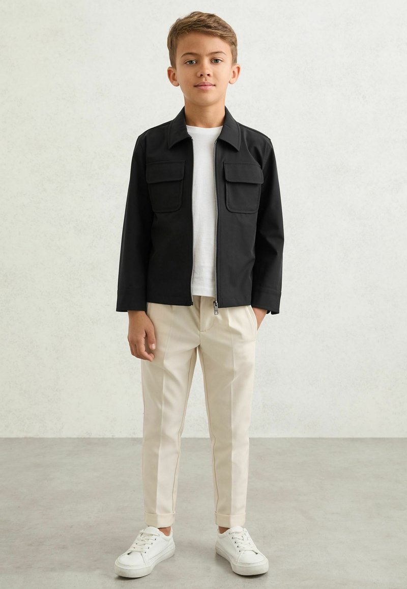 Poika seisoo yksiväristä taustaa vasten, pukeutuneena mustaan vetoketjutakkiin, valkoiseen t-paitaan, beigen värisiin hihdettäviin housuihin ja valkoisiin lenkkareihin.