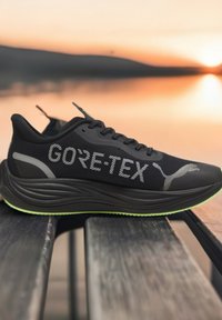 Crne trkaće cipele s Gore-Tex oznakom, gornji dio od teksturirane mreže, lagani dizajn i zeleni akcenti na potplatu. Glatki, aerodinamični oblik.