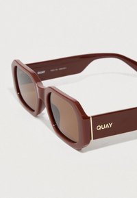 Rudos kvadratinės formos akiniai nuo saulės su storu rėmu, blizgiu paviršiumi ir tamsiais lęšiais. Šonuose yra auksinės spalvos prekinis ženklas.