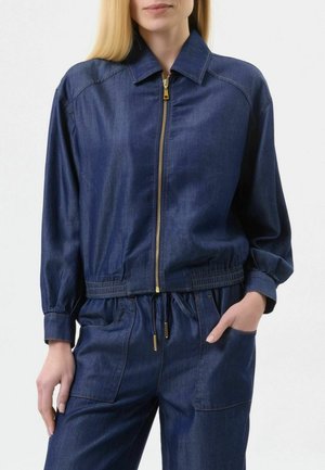 Donna che indossa una giacca di jeans blu scuro con cerniera dorata e pantaloni di jeans a vita alta abbinati, con una mano in tasca, in piedi davanti a uno sfondo bianco.