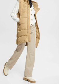 Gilet imbottito beige con colletto alto, sbottonato sopra una maglietta bianca a coste, abbinato a pantaloni beige in corduroy a gamba larga e scarpe di colore chiaro.