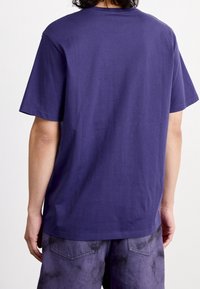 T-shirt a maniche corte in cotone viola scuro, con scollo rotondo e vestibilità ampia. La vista posteriore mostra una texture liscia e un design semplice.