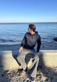Fiatal férfi sötét kapucnis pulóverben és szürke nadrágban, egy beton korláton ülve egy homokos tengerpart mellett, nyugodt óceánra nézve tiszta kék ég alatt.