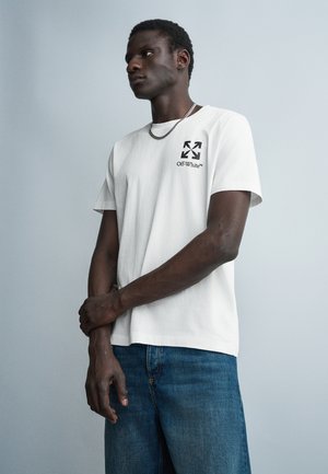 Νέος άνδρας που φοράει λευκό t-shirt Off-White και μπλε τζιν παντελόνι, στέκεται με σταυρωμένα τα χέρια μπροστά σε ένα απλό ανοιχτό γκρι φόντο.