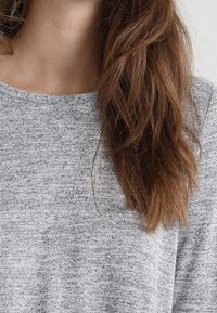 Kvinna med långt brunt hår som bär en ljusgrå texturerad stickad tröja, visad från halsen till midjan mot en enkel bakgrund.