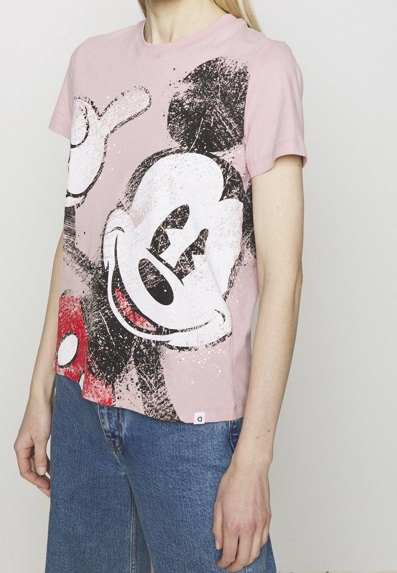 Persona vistiendo una camiseta rosa con un gráfico de Mickey Mouse, combinada con jeans de denim azul, de pie contra un fondo sencillo.