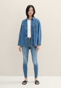 Világoskék farmerring, gombos kialakítással és két mellzsebbel. Fehér póló alatta, hozzá illeszkedő világoskék farmernadrággal és fekete szandálokkal.