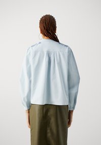 Blusa a righe azzurra chiara con maniche a sbuffo, caratterizzata da dettagli floreali ricamati sul retro e uno yoke plissettato. Abbinata a una gonna verde.