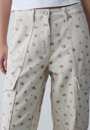 Beige Cargo-Hose mit kleinem rotem Blumenmuster, vorne mit Knopf- und Reißverschluss, seitlichen Taschen mit Klappen, getragen zu einem weißen Oberteil.