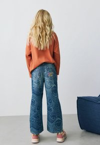 Blugi cu picioare largi, de culoare albastră, cu un model texturat de fețe zâmbitoare și spirale, având o aplicație colorată cu "Smiley" pe buzunarul din spate.