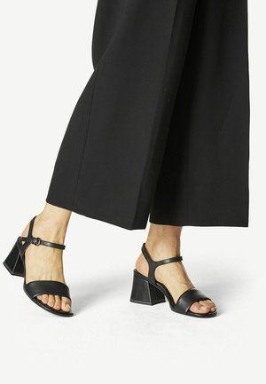 Füße tragen schwarze Sandalen mit offenem Zehenbereich, Blockabsatz und Knöchelriemen, gezeigt mit schwarzen, weit geschnittenen Hosen vor neutralem Hintergrund.
