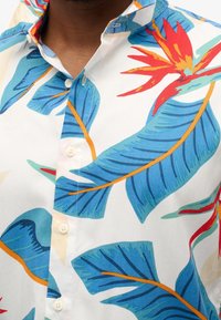 Camisa de manga corta con fondo blanco y grandes patrones de hojas y flores tropicales en azul, verde, rojo y naranja; cuello abotonado.