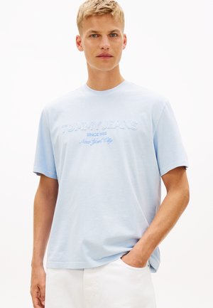 Mladý muž nosiaci svetlomodré tričko Tommy Jeans s nápisom "Since 1985 New York City", stojaci s jednou rukou v bielom vrecku na nohaviciach.