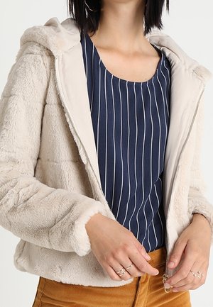 Lys beige pelsjakke med hætte, quiltet design og lynlås. Båret over en marineblå stribet top, der viser en blød tekstur.