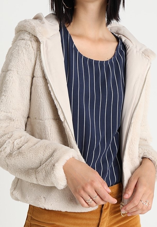 Lys beige pelsjakke med hætte, quiltet design og lynlås. Båret over en marineblå stribet top, der viser en blød tekstur.