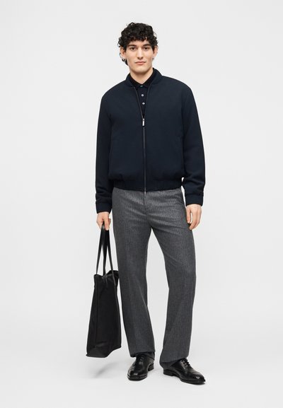 Άνδρας που φοράει μαύρο μπουφάν τύπου bomber, μαύρο πόλο, γκρι παντελόνι και μαύρα παπούτσια, κρατώντας μια μαύρη τσάντα χειρός, στέκεται μπροστά σε λευκό φόντο.