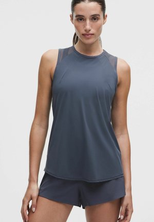 Frau trägt ärmelloses dunkelblaues Sporttop und passende Shorts, schaut mit neutralem Gesichtsausdruck nach vorne vor weißem Hintergrund.