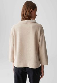 Donna con capelli lunghi che indossa un maglione largo beige a maniche lunghe e pantaloni neri, con la schiena rivolta verso un semplice sfondo grigio.
