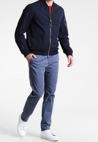 Námornícká bomber bunda se zipem, žebrovanými manžetami a lemem, spárovaná se světle modrými chinos kalhotami a bílými teniskami, což vytváří ležérní vzhled.