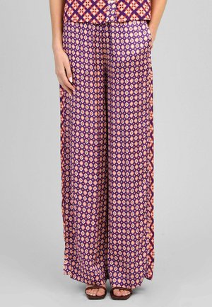 Femme portant un pantalon taille haute à jambes larges et un haut assorti avec des motifs floraux et géométriques violets, roses et oranges, mains partiellement dans les poches.