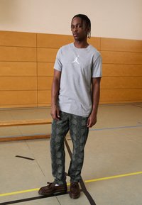 Grå kortærmet t-shirt med et hvidt logo, mønstret mørkegrønne bukser der minder om slangaskin, og brune sko med snørebåndsdetaljer.