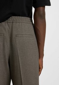 Brown textured trousers feature an elastic waistband and two back pockets, displayed against a black top.