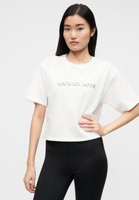 T-shirt branca cropped de algodão, com mangas curtas e logótipo "MICHAEL KORS" em relevo na frente. Combinada com leggings pretas.