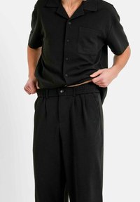 Chemise noire à manches courtes avec des boutons et une poche poitrine, associée à un pantalon plissé assorti avec une taille élastique et un détail logo.