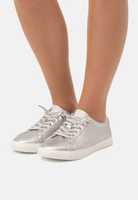 Zapatillas metálicas plateadas con un acabado suave, suela de goma blanca y diseño de cordones. Presentan un logo en el lateral y un cuello acolchado.
