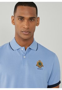 Polo azzurro chiaro in un tessuto texturizzato, con colletto nero e logo ricamato in oro e blu navy. Tre bottoni al collo.