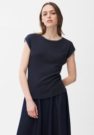 Młoda kobieta ubrana w dopasowany granatowy top z krótkim rękawem z białym wykończeniem oraz ciemną plisowaną spódnicę, stojąca na białym tle.