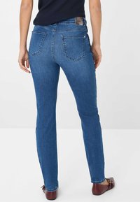 Donna che indossa jeans slim-fit blu medio con tasche posteriori e scarpe slip-on marroni, in piedi davanti a uno sfondo chiaro e uniforme.