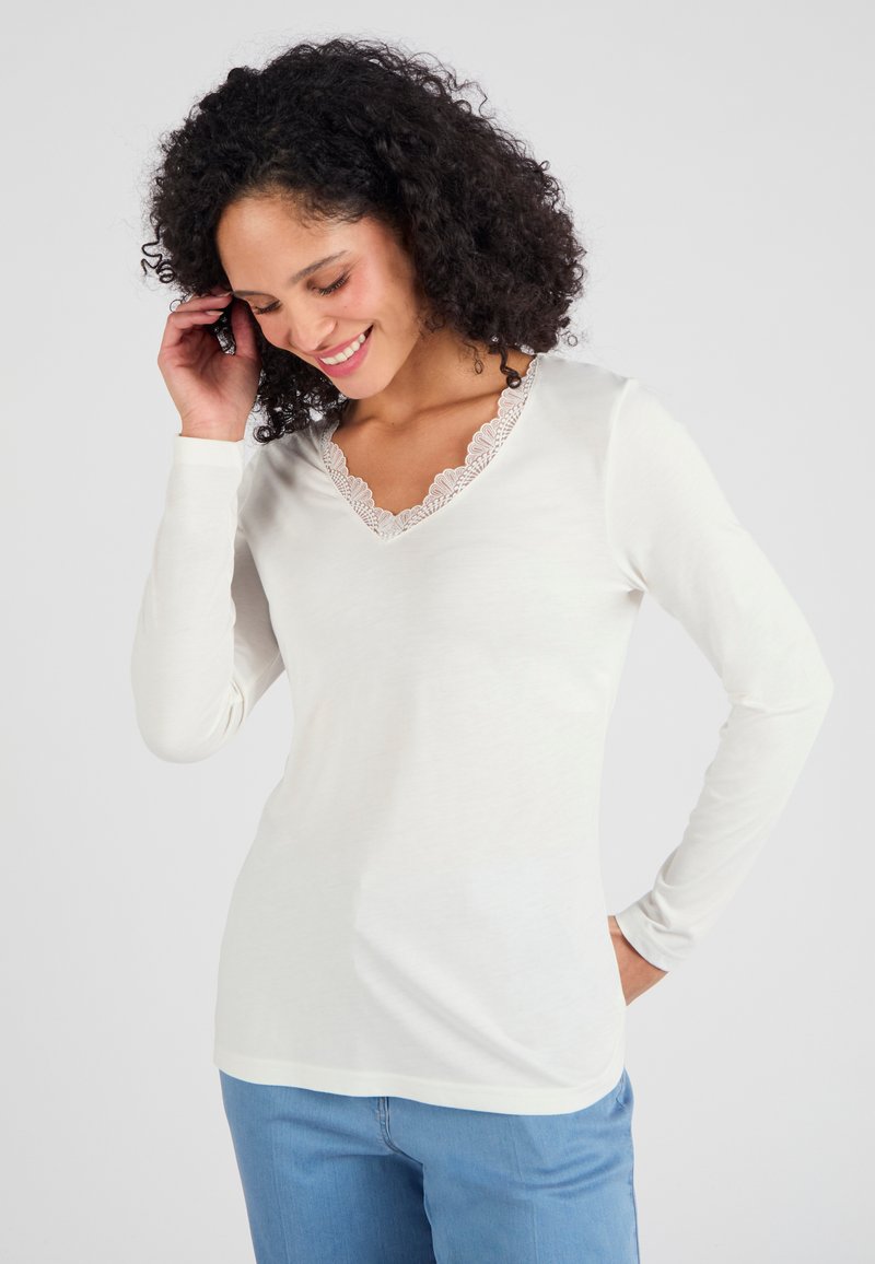 Wit lange mouwen top met een V-hals en kantafwerking, gemaakt van zachte stof, met een getailleerde pasvorm en afgeronde zoom. G gedragen met lichtblauwe jeans.